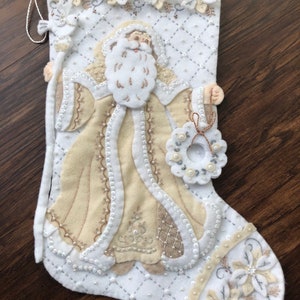 Puede incluir: Calcet&iacute;n de Navidad de fieltro blanco con una figura de Pap&aacute; Noel beige que sostiene una corona. El calcet&iacute;n est&aacute; decorado con cuentas y flores blancas y beige.
