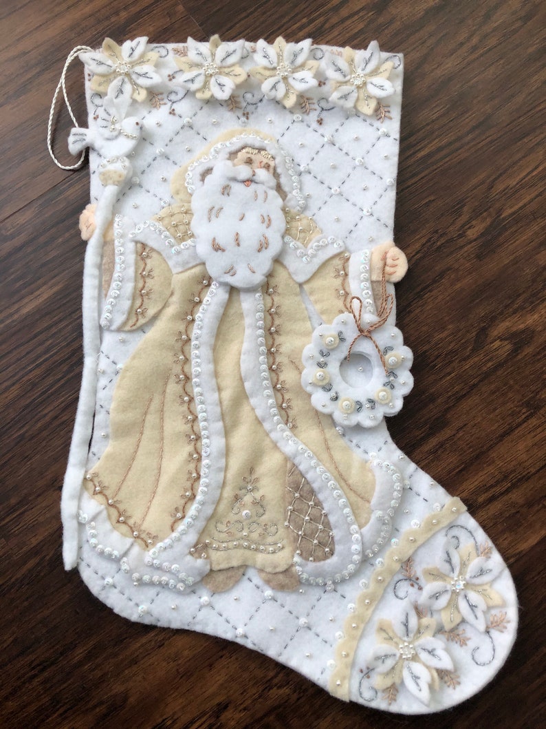 Puede incluir: Calcet&iacute;n de Navidad de fieltro blanco con una figura de Pap&aacute; Noel beige que sostiene una corona. El calcet&iacute;n est&aacute; decorado con cuentas y flores blancas y beige.