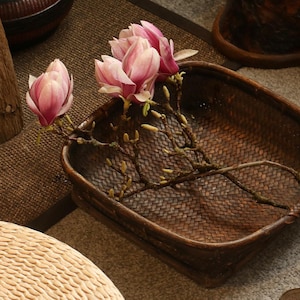 May include: A brown woven basket displays pink magnolia flowers and branches. The basket is on a grey surface, with a wooden stool and a woven seat in the background. Metal pliers are also visible.