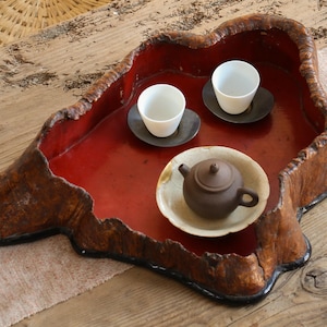 Puede incluir: Bandeja de madera lacada en rojo, hecha a mano, con una forma orgánica. Contiene dos tazas de té blancas sobre platillos oscuros y una pequeña tetera marrón sobre un plato crema, ideal para servir té.