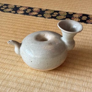 May include: A gray ceramic teapot with a unique spout design. The teapot has a round body and a small, round opening on top.
