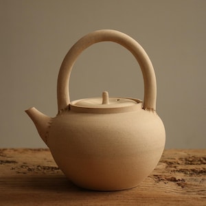 May include: A beige ceramic teapot with a rounded body, a curved handle, and a small spout. The lid has a small knob. The teapot is sitting on a wooden surface.