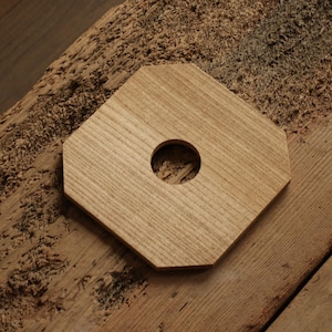 Puede incluir: Un posavasos octogonal de madera clara con un agujero central, sobre una superficie de madera envejecida. El posavasos tiene vetas de madera visibles. El fondo incluye más madera y una textura tejida.