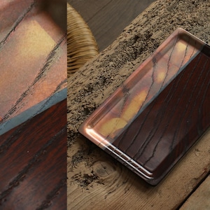 Può includere: Vassoio rettangolare con un design bicolore. Una metà presenta un motivo a venature del legno marrone rossiccio caldo, mentre l'altra ha una miscela di tonalità rame, oro e nero. Il vassoio ha una finitura lucida.