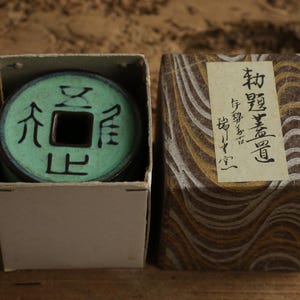 Op de afbeelding: Een turquoise keramisch muntvormig object met zwarte Japanse karakters wordt getoond in een witte doos. Daarnaast een decoratieve doos met een label met Japanse tekst. De dozen staan op een houten oppervlak.