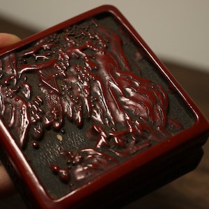 May include: A red lacquered box with a carved design of a mountain scene. The box is made of wood and has a dark brown interior.