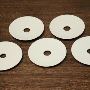 May include: Five white ceramic coasters with a circular hole in the center. The coasters are arranged in a semi-circle on a wooden surface.
