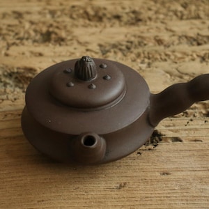 Könnte beinhalten: Eine braune Keramik-Teekanne mit rundem Körper und gebogenem Griff. Der Deckel hat einen kleinen Knopf und mehrere kleine dekorative Noppen. Der Ausguss ist kurz und abgewinkelt. Die Teekanne wird wahrscheinlich zum Brühen von Tee verwendet.