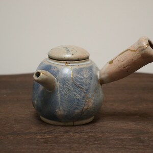 Op de afbeelding: Een klein, blauw en wit aardewerken theepotje met een gouden craquelé-glazuur en een roze handvat. De theepot heeft een rond lichaam en een tuit aan de zijkant.