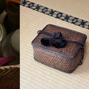 May include: A brown woven basket with black tassels and a black strap. The basket is sitting on a straw mat with a black and gold patterned border.