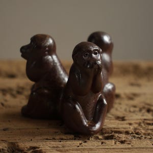 Op de afbeelding: Drie bruine apenbeeldjes in zittende positie. Eén aap bedekt zijn mond met zijn handen, een ander bedekt zijn oren en de derde bedekt zijn ogen. De beeldjes staan op een houten oppervlak.