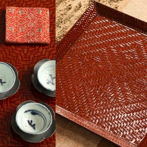 May include: A still-life arrangement featuring a red woven tray, small white and blue teacups with saucers, a red patterned cloth, and a white teapot on a metal tray. The items are arranged on a red woven surface.