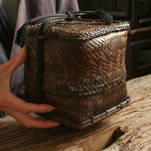 May include: A square, woven, brown basket with a black handle and tassels. The basket is made of natural materials and has a rustic, antique look.