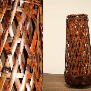 Puede incluir: Una cesta de bambú tejida marrón con un patrón de diamante. La cesta es alta y cilíndrica.