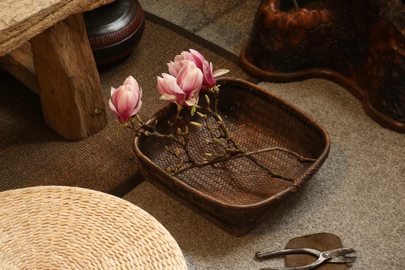May include: A brown woven basket displays pink magnolia flowers and branches. The basket is on a grey surface, with a wooden stool and a woven seat in the background. Metal pliers are also visible.