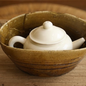 May include: A small white ceramic teapot with a round lid and handle sits inside a larger, deep, brown-toned ceramic bowl. The bowl features a visible crack along its upper rim and a textured, earthy finish, contrasting with the smooth white teapot.