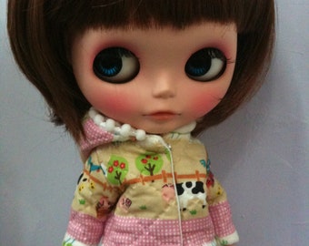 May include: A doll with brown hair wearing a pink and beige jacket with a farm animal print and a pink polka dot skirt.