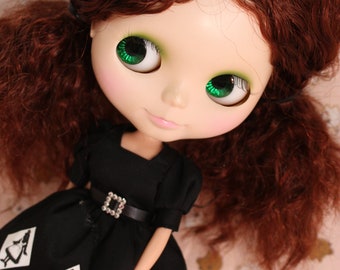 May include: A doll with long, curly auburn hair wearing a black dress with white accents. The dress has a black belt with a silver buckle and a ruffled hem. The doll has green eyes and a painted smile.