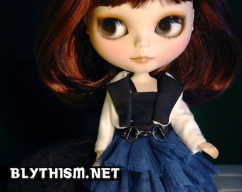 May include: A doll with long, dark red hair, wearing a black and white waistcoat over a blue tulle skirt. The doll has large, blue eyes and a sweet expression. The doll is wearing a black belt with silver buckles. The doll is standing on a black surface. The text "BLYTHISM.NET" is visible in the lower left corner of the image.