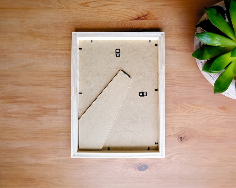 May include: A rectangular, light-colored wooden picture frame, viewed from the back, with a cardboard backing and a built-in easel. The frame is set on a light brown wooden surface, with a green succulent plant in a white pot in the background.