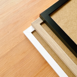 May include: Three picture frames in black, white, and light wood tones are arranged diagonally on a light wood surface. The frames have a simple, modern design, with a neutral-colored backing visible within the frames.