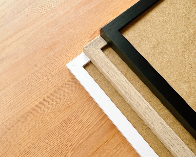 May include: Three picture frames in black, white, and light wood tones are arranged diagonally on a light wood surface. The frames have a simple, modern design, with a neutral-colored backing visible within the frames.