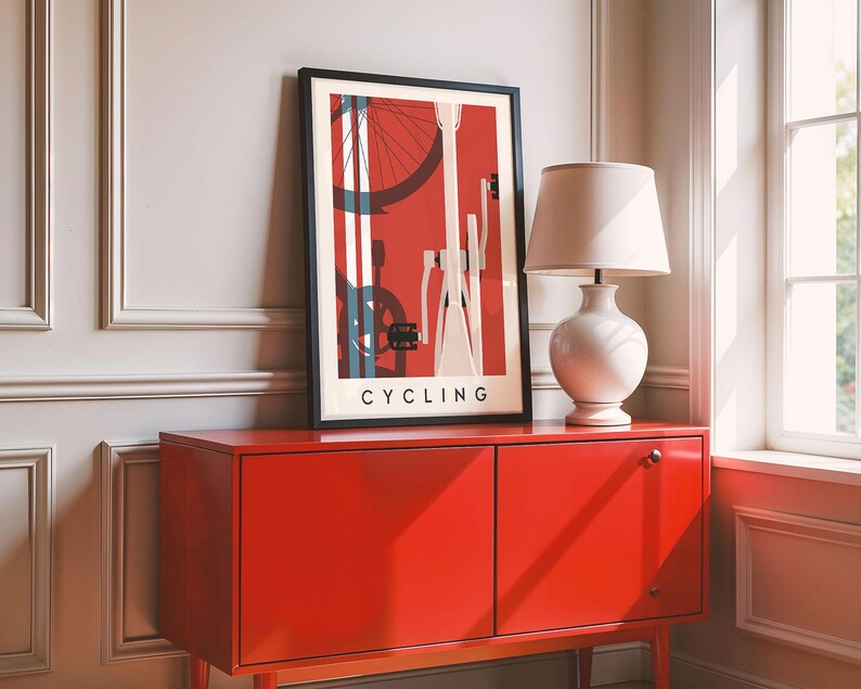 May include: A framed art print with a cycling theme, featuring a red background and bicycle components. The print is displayed on a bright red cabinet, next to a white lamp with a beige lampshade. The word "CYCLING" is printed at the bottom of the artwork.
