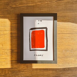 May include: A framed art print with a bold red square and black border on a white background. The text "I AM A FRAME" is handwritten in black. The frame is black and sits on a wooden surface.