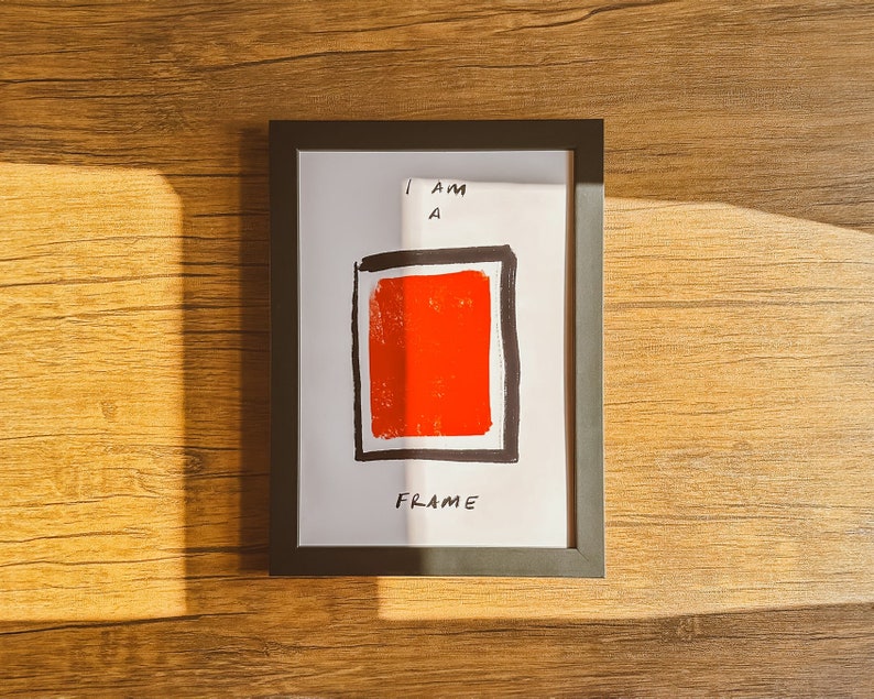May include: A framed art print with a bold red square and black border on a white background. The text "I AM A FRAME" is handwritten in black. The frame is black and sits on a wooden surface.