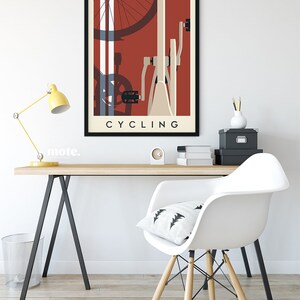 May include: A framed art print featuring a stylized bicycle illustration in shades of red, cream, and blue. The word "CYCLING" is printed in a bold, sans-serif font at the bottom. The artwork is displayed on a white wall above a wooden desk.
