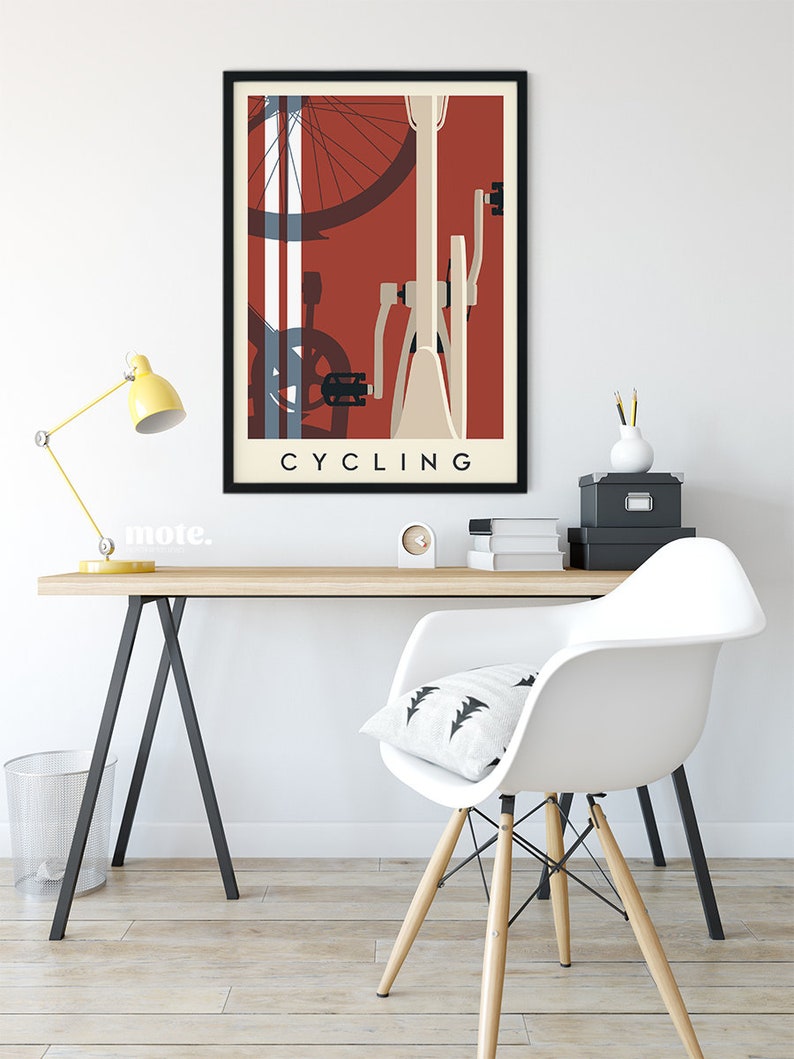 May include: A framed art print featuring a stylized bicycle illustration in shades of red, cream, and blue. The word "CYCLING" is printed in a bold, sans-serif font at the bottom. The artwork is displayed on a white wall above a wooden desk.