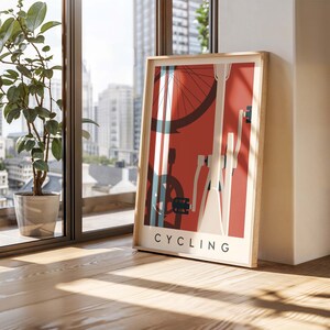 May include: A framed poster with a cycling theme. The artwork features a bicycle wheel, frame, and pedals in white, blue, and black against a red background. The word "CYCLING" is printed in white at the bottom. The frame is made of light-colored wood.