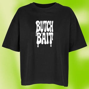 Könnte beinhalten: Ein schwarzes T-Shirt mit kurzen Ärmeln und dem weißen Schriftzug BUTCH BAIT, der in einer stilisierten, tropfenden Schriftart mittig auf der Brust aufgedruckt ist. Das Kleidungsstück hat einen Rundhalsausschnitt und eine lockere Passform.