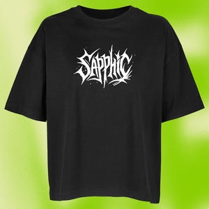 Könnte beinhalten: Schwarzes T-Shirt mit dem Wort "Sapphic" in weißer, stilisierter Schrift. Der Text hat ein gotisches, stacheliges Design. Das Shirt hat kurze Ärmel und einen Rundhalsausschnitt. Der Hintergrund ist ein Grün-Gelb-Verlauf.