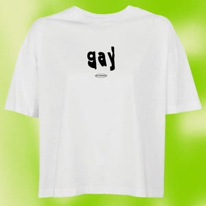 Könnte beinhalten: Weißes, bauchfreies T-Shirt mit dem Wort "gay" in schwarzer, stilisierter Schrift. Das Wort ist in einer leicht verzerrten Schriftart.