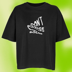 Könnte beinhalten: Ein schwarzes T-Shirt mit kurzen Ärmeln und weißem Text auf der Brust, der DON'T JUDGE BODIES lautet. Die Schrift ist in einem gestapelten, leicht verzerrten und wellenförmigen Grafikdesign in der Mitte des Kleidungsstücks angeordnet.