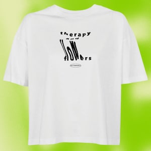 Könnte beinhalten: Weißes T-Shirt mit schwarzem Text, der in einer verzerrten Schriftart "Therapie und Blumen" lautet. Der Text ist mittig auf dem Shirt platziert. Das Shirt hat eine lockere Passform.