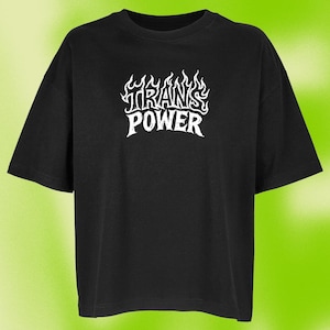 Könnte beinhalten: Schwarzes T-Shirt mit dem weißen Text "TRANS POWER" in einer flammenartigen Schriftart. Das Shirt hat einen Rundhalsausschnitt und eine lockere Passform. Der Hintergrund ist ein Farbverlauf aus Grün und Gelb.