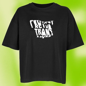 Könnte beinhalten: Ein schwarzes T-Shirt mit kurzen Ärmeln und einem weißen Grafikdruck auf der Brust. Das Design besteht aus dem Text "PROTECT TRANS LIVES" in einer verzerrten, wellenförmigen Schriftart. Das Kleidungsstück hat einen Rundhalsausschnitt und eine lockere Passform.