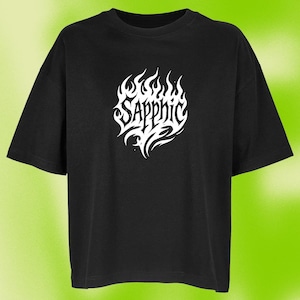Könnte beinhalten: Schwarzes T-Shirt mit einem weißen, stilisierten "Sapphic"-Design in einer flammenartigen Schriftart. Das Shirt hat kurze Ärmel und eine lockere Passform. Der Hintergrund ist ein grüner Farbverlauf.