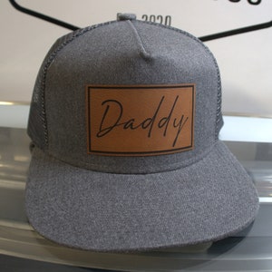 Può includere: Cappellino da camionista grigio e marrone con una toppa in pelle marrone che dice "Daddy" in corsivo.