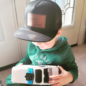 Peut inclure: Un chapeau de camionneur en maille noire avec un patch en cuir marron qui dit "Brody James". Le chapeau est porté par un enfant portant un sweat à capuche vert avec les mots "Old Navy" sur le devant.