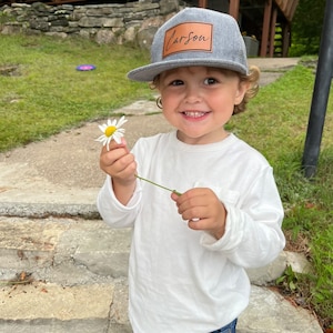 Peut inclure: Un jeune enfant portant une casquette de baseball grise et marron avec le nom "Carson" écrit dessus. L'enfant tient une marguerite blanche et sourit à la caméra.