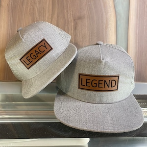 Può includere: Due cappellini da camionista in rete grigia con toppe in pelle marrone. La toppa sul cappello a sinistra dice "LEGACY" e la toppa sul cappello a destra dice "LEGEND".