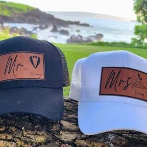 Op de afbeelding: Twee truckerpetten, één zwart en één wit, met bruine leren patches. De zwarte pet heeft "Mr." en een smoking-afbeelding, terwijl de witte pet "Mrs." zegt met een jurkafbeelding. Beide hebben de naam "Irving" eronder.