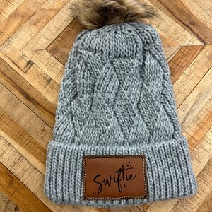 Op de afbeelding: Grijze gebreide beanie met een bruine nep bont pompon en een bruin leren patch met de tekst "Swiftie" met een glitterontwerp.