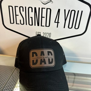 Peut inclure: Casquette de camionneur noire avec un patch en cuir marron portant l'inscription "DAD" en lettres grasses, avec "adelinn & everly" en cursive en dessous. Le chapeau a un dos en filet et une visière incurvée. Le fond présente le texte "DESIGNED 4 YOU" et une illustration de chapeau.