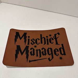 Può includere: Tappetino per mouse in pelle marrone con testo nero che recita "Mischief Managed" con una grafica di bacchetta.