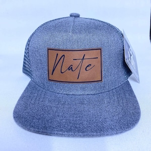 May include: A gray denim trucker hat with a brown leather patch that says "Nate" in black script.