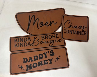 May include: Four brown leather patches with black text. The patches read "Moen", "Kinda Kinda Broke Bougie", "Daddy's Money", and "Chaos Container".